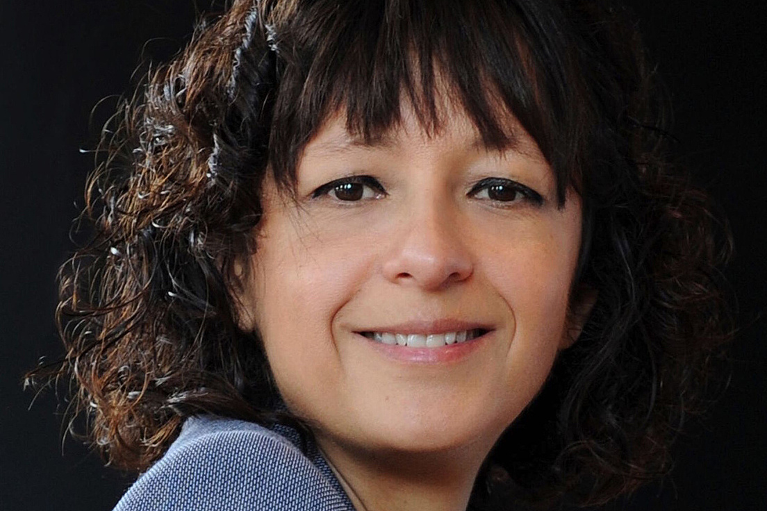Nobelpreis für Chemie Emmanuelle Charpentier blickt in die Kamera, der Hintergrund ist dunkel, sie trägt eine blaues Oberteil. Ihre Haare sind mittellang, braun und gelockt.