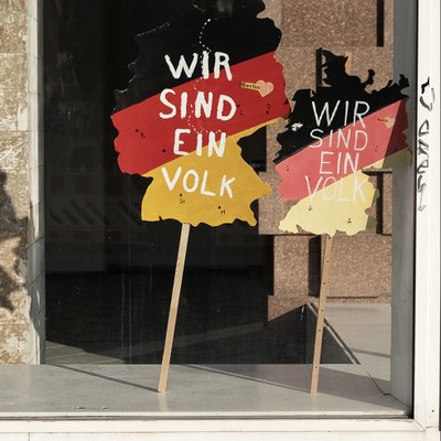 Zwei Platten die an jeweils einem Stock befestigt sind stehen in einem Schaufenster.  Sie haben die Form des Deutschland-Umrisses und in den Farben der Deutschen Flagge. Auf diesen steht "Wir sind ein Volk".