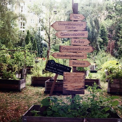 An einem Stcok sind Wegweiser, die in einem urbanen Garten in verschiedenen Richtungen zeigen. Beschriftet mit u.a. Bienen, Acker, Gastronomie.