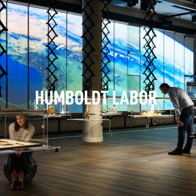 Im Vordergrund steht „Humboldt Labor". Dahinter schauen Besucher:innen in die hängenden Vitrinen im Humboldt Labor. Im Hintegrund ist die interaktive Forschungswand.