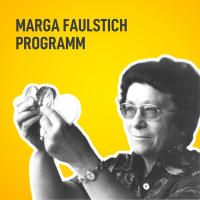 Eine ältere Frau, Marga Faulstich, mit kurzen Haaren, Brille und einer Bluse mit Mustern hält eine Glaslinse hoch und sieht sie prüfend an. 