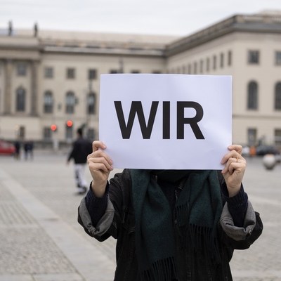 Eine Person hält ein großes Blatt Papier vor ihrem Gesicht hoch. Darauf steht in Großbuchstaben das Wort WIR. Im Hintegrund ist das Hauptgebäude der Humboldt-Universität zu sehen.