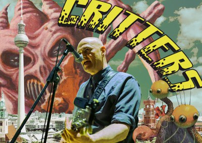 Critters Kevin Mooney mit umgehängter Gitarre vor dem Critters Plakat