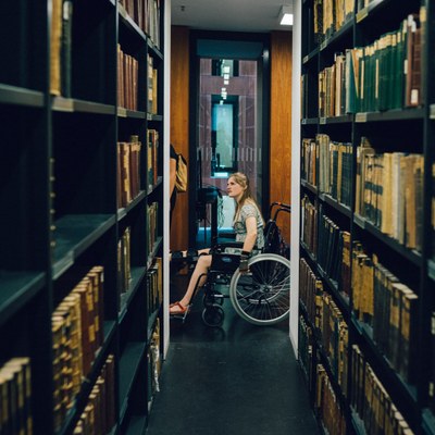 Blick durch zwei historische Bücherregale des Grimmzentrums auf eine sich umschauende Studentin im Rollstuhl.
