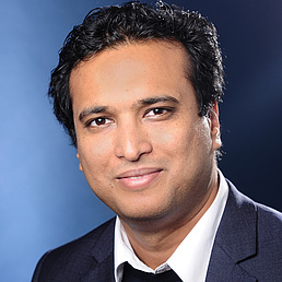 Kallol Ray hat ein weißes Hemd, einen dunkelfarbenen Pullover und einen schwarzen Anzug an. Er blickt freundlich in die Kamera.