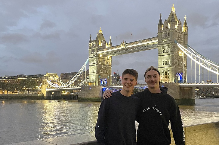 Zwei Personen stehen vor der beleuchteten Tower Bridge in London am Abend mit Fluss im Vordergrund