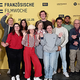 Studierende und Xavier Bihan posieren in einem Gruppenfoto vor einem gelben Plakat der Französischen Filmwoche. Sie tragen alle bequeme und lockere Kleidung und blicken fröhlich in die Kamera.