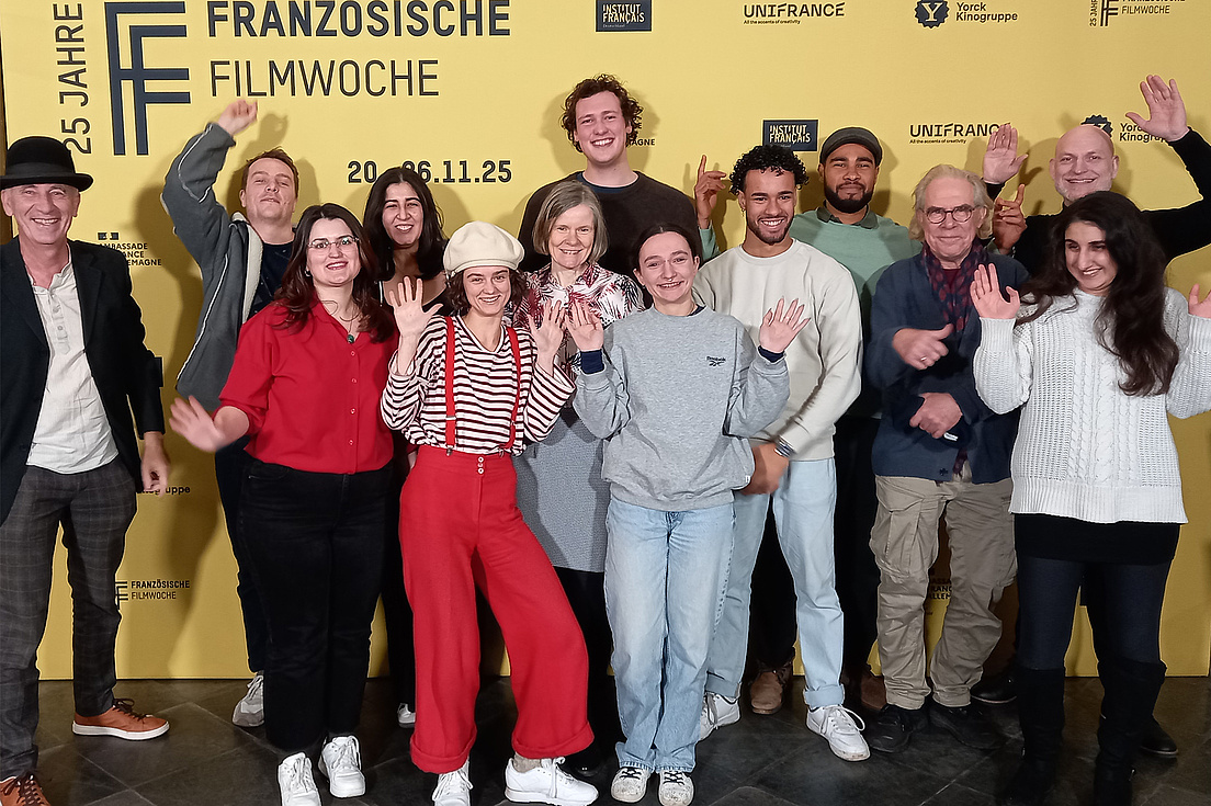 Gruppenfoto der Studierenden und Xavier Bihan Studierende und Xavier Bihan posieren in einem Gruppenfoto vor einem gelben Plakat der Französischen Filmwoche. Sie tragen alle bequeme und lockere Kleidung und blicken fröhlich in die Kamera.
