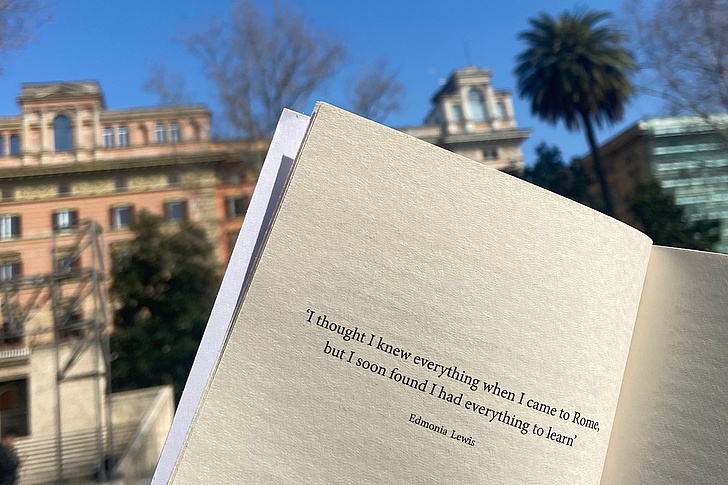Offenes Buch mit Zitat „I hate everything when I leave Rome, but I found everything when I leave Rome“ von E. M. Forster vor römischen Gebäuden und Palmen bei blauem Himmel.