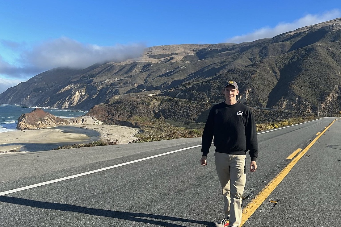 Person in schwarzem Pullover und beiger Hose geht auf einer Straße mit gelber Mittellinie entlang, im Hintergrund Hügel und Küstenlandschaft