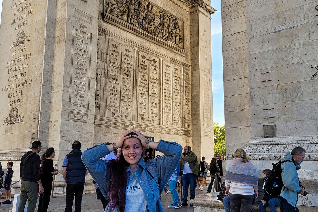 Freudestrahlende junge Frau mit langen lila Strähnen in Jeansjacke hält überrascht die Hände an den Kopf vor dem Arc de Triomphe mit Namenslisten und Touristen bei blauem Himmel. ​