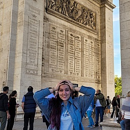 Freudestrahlende junge Frau mit langen lila Strähnen in Jeansjacke hält überrascht die Hände an den Kopf vor dem Arc de Triomphe mit Namenslisten und Touristen bei blauem Himmel. ​
