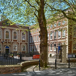 Historische Backsteingebäude im Innenhof einer Universität, mit roten Banner "King's College London" an den Fassaden, umgeben von hohen Bäumen mit grünem Laub, sonniges Wetter und schmiedeeisernem Zaun.