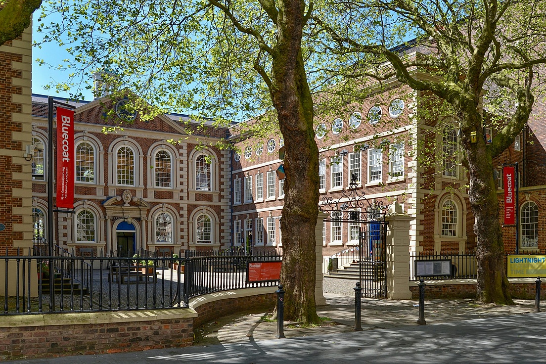 Historische Backsteingebäude im Innenhof einer Universität, mit roten Banner "King's College London" an den Fassaden, umgeben von hohen Bäumen mit grünem Laub, sonniges Wetter und schmiedeeisernem Zaun.