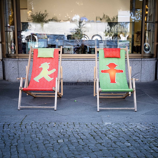 Zwei Liegestühle vor einem Geschäft. Links ein roter Liegestuhl mit grünem Ampelmännchen und rechts ein grüner mit rotem Ampelmännchen.