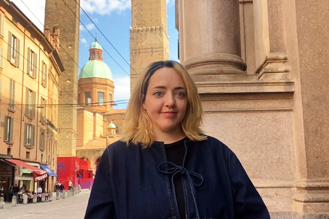 Person mit schulterlangen blonden Haaren und dunkler Jacke steht vor einer historischen Säule in einer Stadt mit sichtbaren Türmen und Kuppeln im Hintergrund