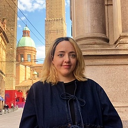 Person with shoulder-length blonde hair and dark jacket standing in front of a historic column in a city with visible towers and domes in the background