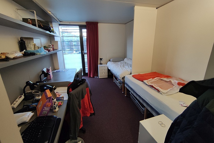 Studentisches Zimmer mit Schreibtisch, Bett, Bücherregal, rotem Vorhang und Fenster zum Grünbereich