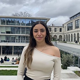Young woman with long hair and one-shoulder top standing in front of a modern building labeled 'Sciences Po' with several people sitting on stairs.