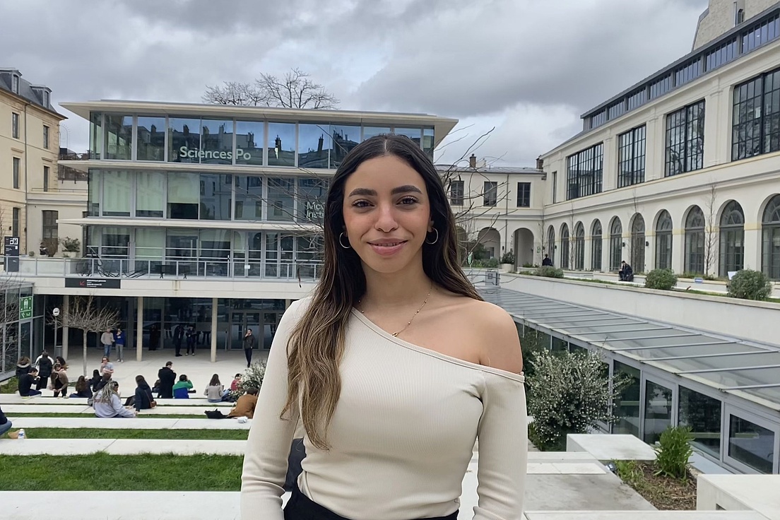 Junge Frau mit langem Haar und offener Schulter steht vor einem modernen Gebäude mit der Aufschrift 'Sciences Po' und mehreren Personen auf einer Treppe.