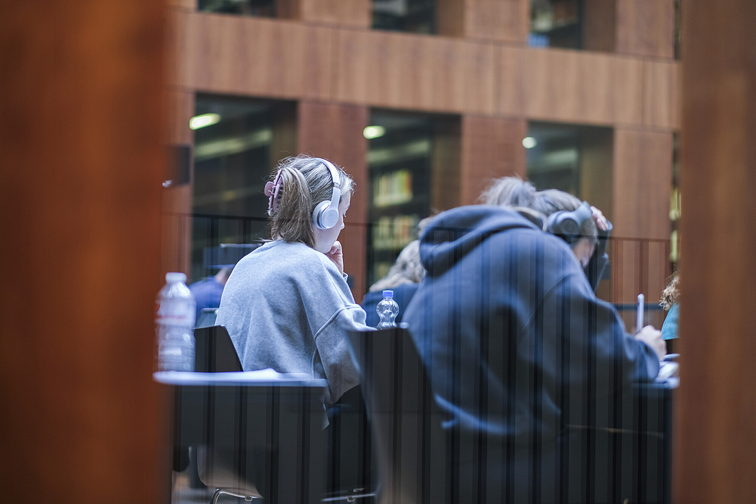 Blick durch eine Glastür des Grimm-Zentrums der HU. Studierende sitzen an Schreibtischen des großen Lesesaals und arbeiten.