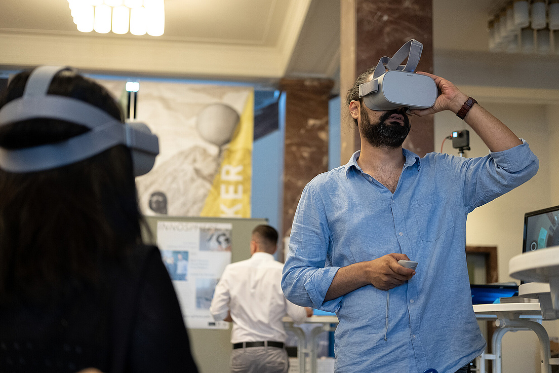 A man in a room wearing a VR headset. In the foreground, you can see the back of the head of a person with long hair, who is also wearing a VR headset.