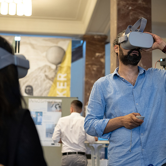 Ein Mann in einem Raum, der eine VR-Brille trägt. Im Vordergrund sieht man den Hinterkopf einer Person mit langen Haaren, die ebenfalls eine VR-Brille trägt.