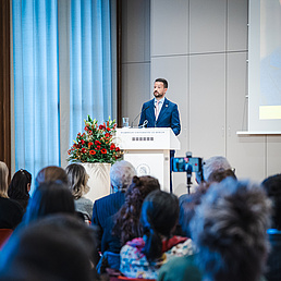 Jacov Milatović wie er am Pult steht und seine Rede hält. Im vorderen Teil des Bildes befindet sich Publikum, das mit dem Rücken zur Kamera sitzt.