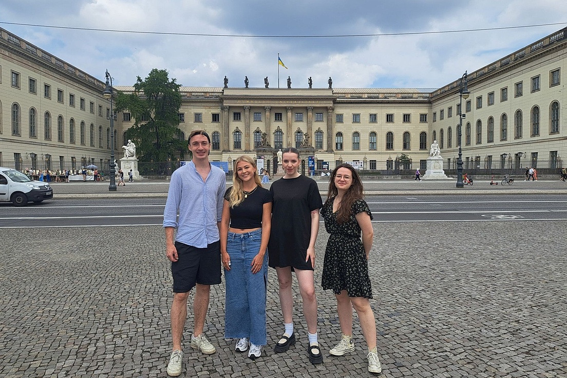 Vier Personen stehen auf einem gepflasterten Platz vor einem großen historischen Gebäude mit Säulen und einer ukrainischen Flagge auf dem Dach.