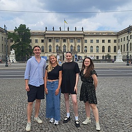 Vier Personen stehen auf einem gepflasterten Platz vor einem großen historischen Gebäude mit Säulen und einer ukrainischen Flagge auf dem Dach.