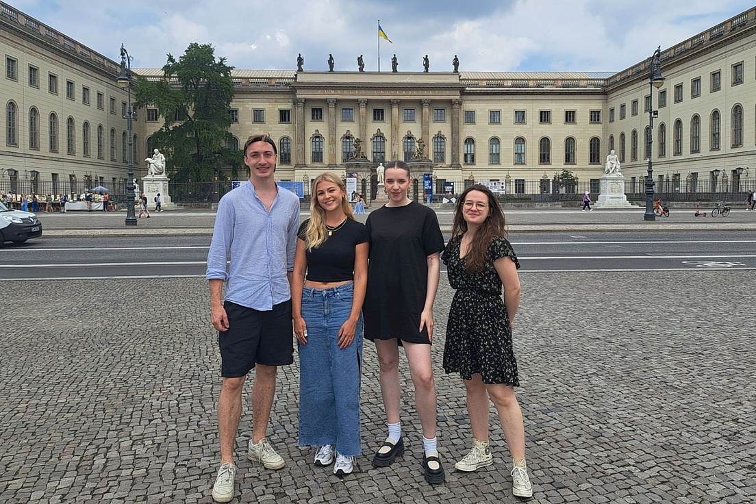 Vier Personen stehen auf einem gepflasterten Platz vor einem großen historischen Gebäude mit Säulen und einer ukrainischen Flagge auf dem Dach.