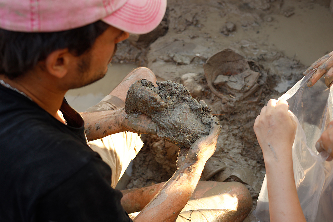 Ein Mann in einer rosa Kappe hält einen mit Schlamm bedeckten Fund, während eine andere Person einen Plastikbeutel hält.