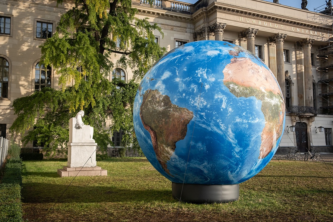 Large globe displayed on a lawn in front of the main building of Humboldt University in Berlin, next to a white statue and surrounded by trees and historic architecture.