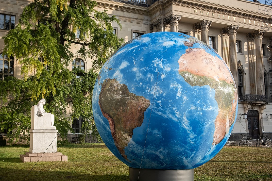 Große Erdkugel auf einer Wiese vor dem Hauptgebäude der Humboldt-Universität in Berlin, neben einer weißen Statue und umgeben von Bäumen und historischer Architektur.
