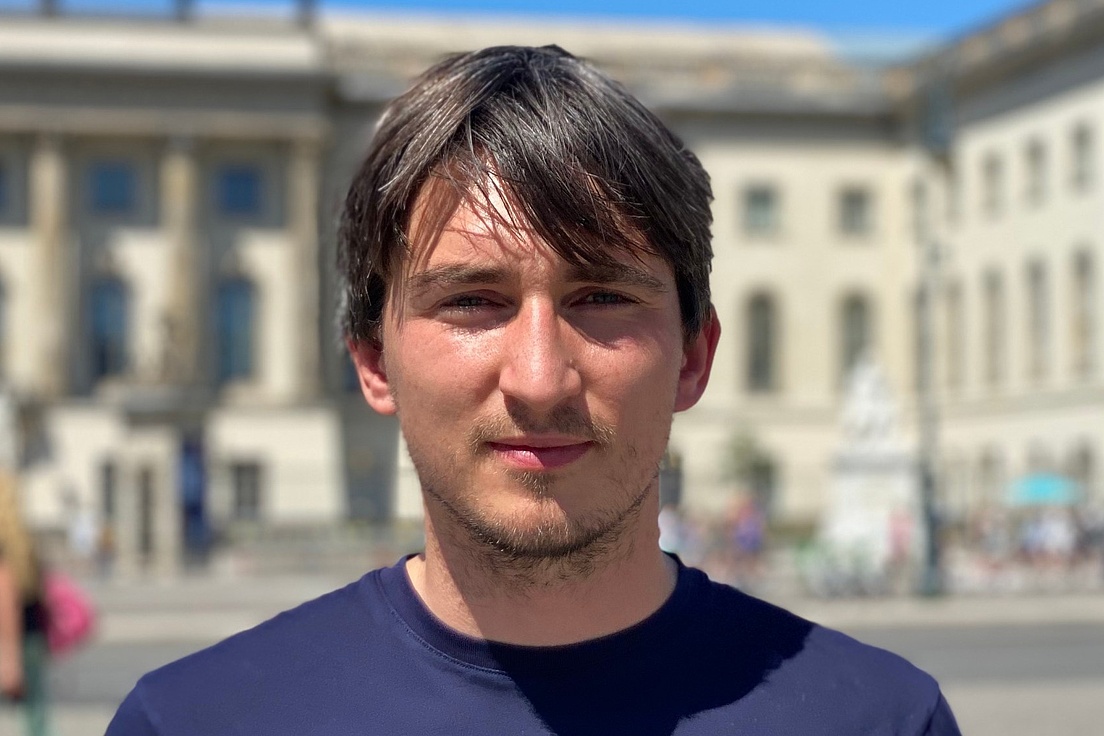 Porträt eines jungen Mannes mit dunklem Haar und Bart in blauem T-Shirt, der vor dem historischen Gebäude mit Säulen und Skulpturen lächelt, Passanten und ukrainische Flagge im Hintergrund.