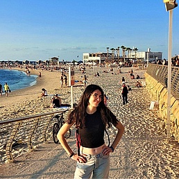 Person mit schwarzem Oberteil und Jeans steht am Strand vor einer Promenade mit Geländer und Menschen im Hintergrund