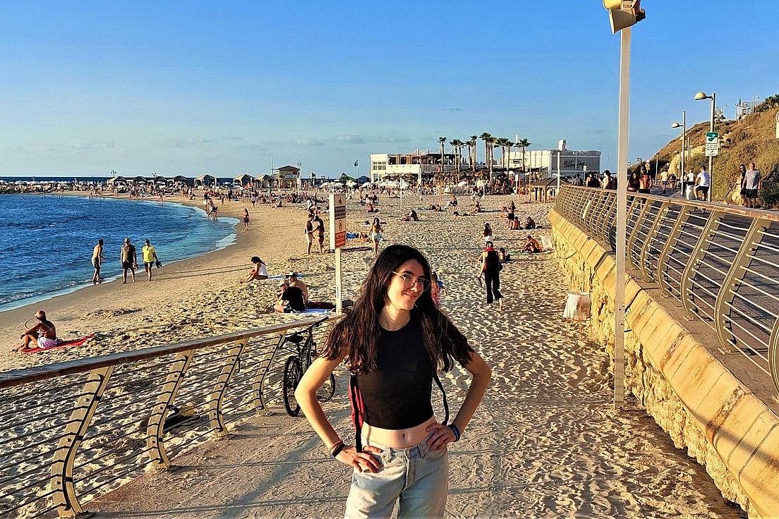 Person mit schwarzem Oberteil und Jeans steht am Strand vor einer Promenade mit Geländer und Menschen im Hintergrund