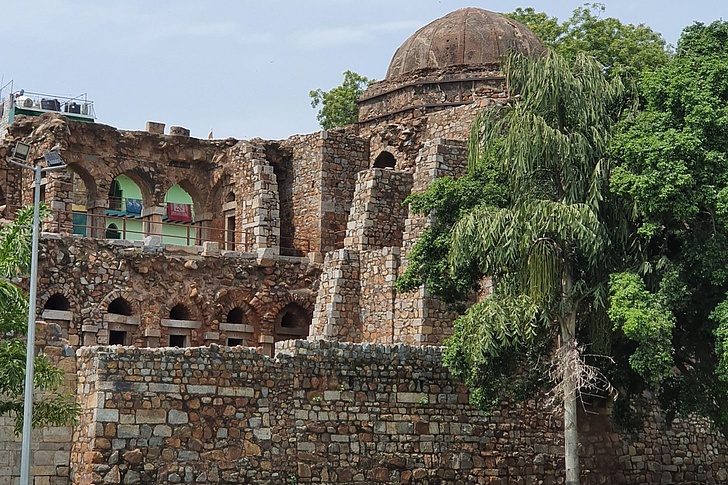 Ruinen des Hauz Khas Fort mit Kuppeldach, Bögen und zerfallenen Sandsteinmauern im Deer Park, umgeben von hängenden Bäumen und Laternen unter klarem Himmel.