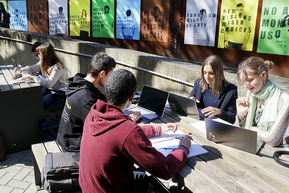 Studierende sitzen an einem langen Holztisch im Freien und arbeiten mit Laptops und Notizbüchern, im Hintergrund sind bunte Plakate an einer Mauer angebracht.