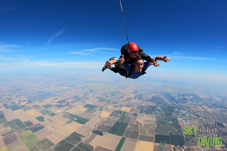 Zwei Personen beim Tandem-Fallschirmsprung über einer landwirtschaftlich geprägten Landschaft unter blauem Himmel