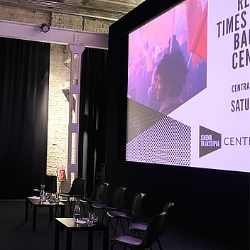 Event room with chairs and two low tables with water for the speakers and, in the background, a slide show projected onto a white screen.