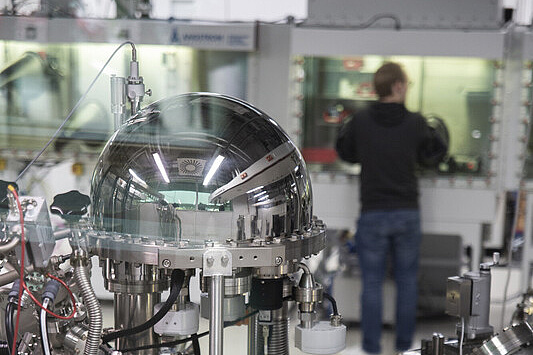 Open-Humboldt-Forschung, Campus Adlershof Das Bild zeigt ein hochkomplexes wissenschaftliches Gerät, vermutlich ein Teil eines Vakuum- oder Hochvakuum-Systems, das in einem Labor eingesetzt wird. Im Vordergrund ist ein glänzender, halbkugelförmiger Behälter zu sehen, der aus Metall gefertigt ist und an mehreren Stellen mit Schläuchen, Kabeln und anderen technischen Komponenten verbunden ist.