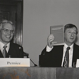 Jean-Claude Juncker sitzt an einem Tisch neben Herr Pernice und hält ein Buch in die Höhe.