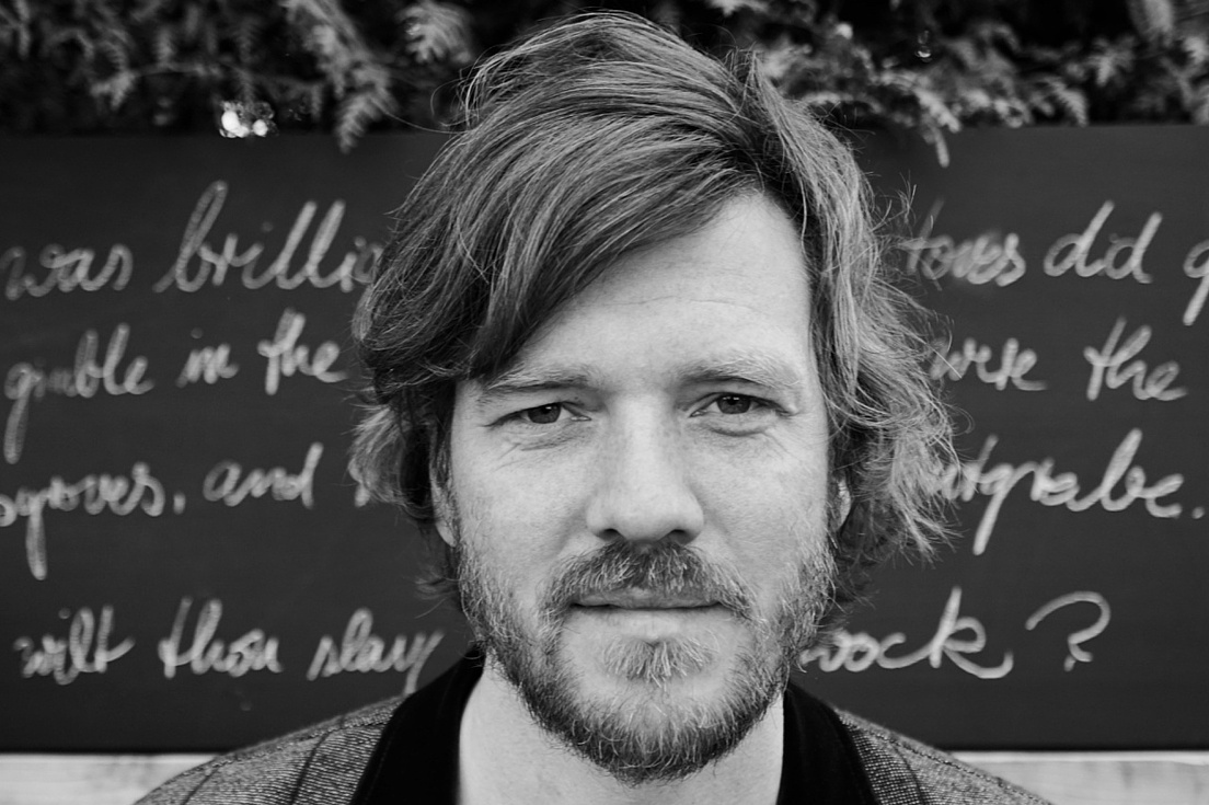 Benjamin Kiesewetter erhält ERC Starting Grant Ein Schwarz-Weiß-Porträt Benjamin Kieswetters. Er hat lockiges, mittellanges Haar und einen Bart mit Schnurrbart. Der Mann blickt direkt in die Kamera mit einem neutralen, leicht nachdenklichen Gesichtsausdruck. Er trägt ein kariertes Jackett mit einem dunklen Kragen über einem weißen T-Shirt.