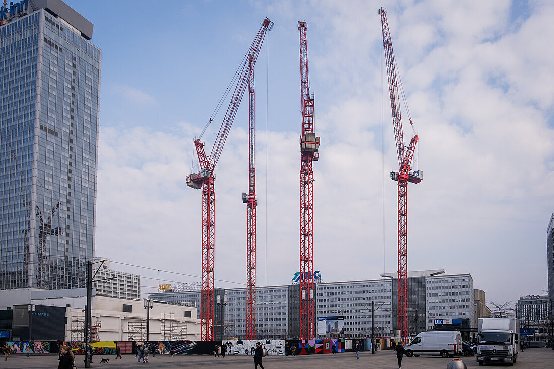 Das Bild zeigt eine städtische Szene mit mehreren Baustellenkranen im Vordergrund. Die Krane sind rot und dominieren das Bild, da sie hoch in den Himmel reichen. Im Hintergrund sind moderne Hochhäuser zu sehen.