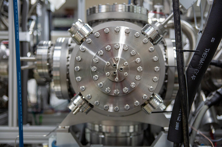 Das Bild zeigt eine Nahaufnahme eines komplexen technischen Geräts, vermutlich ein Teil einer wissenschaftlichen Apparatur oder eines industriellen Maschinenelements. Im Zentrum des Bildes ist eine runde, metallische Struktur mit vielen Schrauben zu sehen, die symmetrisch angeordnet sind und das Gerät zusammenhalten.