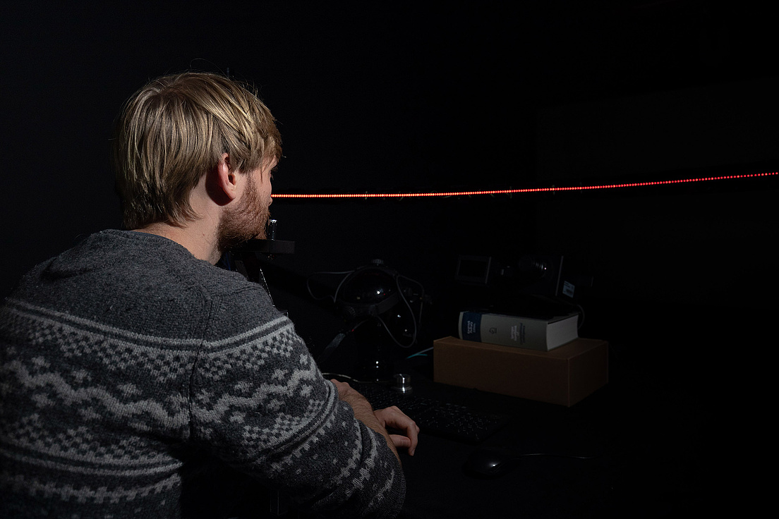 Ein Mann in einem gemusterten Pullover sitzt in einem dunklen Raum und beobachtet eine rote Laserlinie.