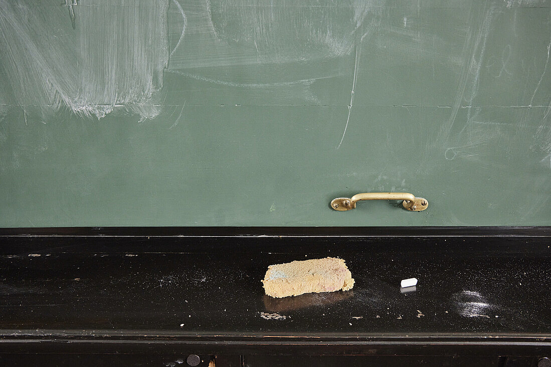 Eine Tafel, vor der ein Schwamm und Reste von Kreide liegen. Die Tafel ist gewischt, Kreidereste hängen noch an ihr. 