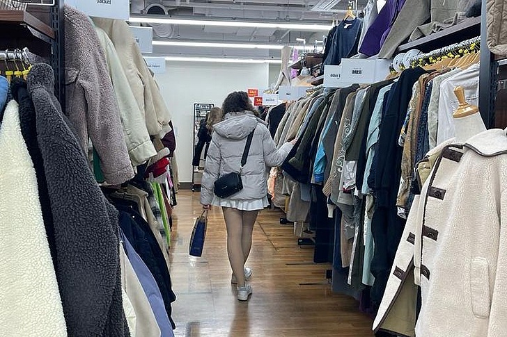 Eine Person mit kurzem Rock und Kapuzenjacke geht durch einen Gang mit beidseitig hängender Kleidung in einem Secondhand-Laden