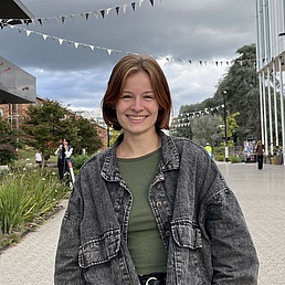 Person with shoulder-length hair wearing green top and black denim jacket standing in an urban plaza with modern buildings and trees in the background